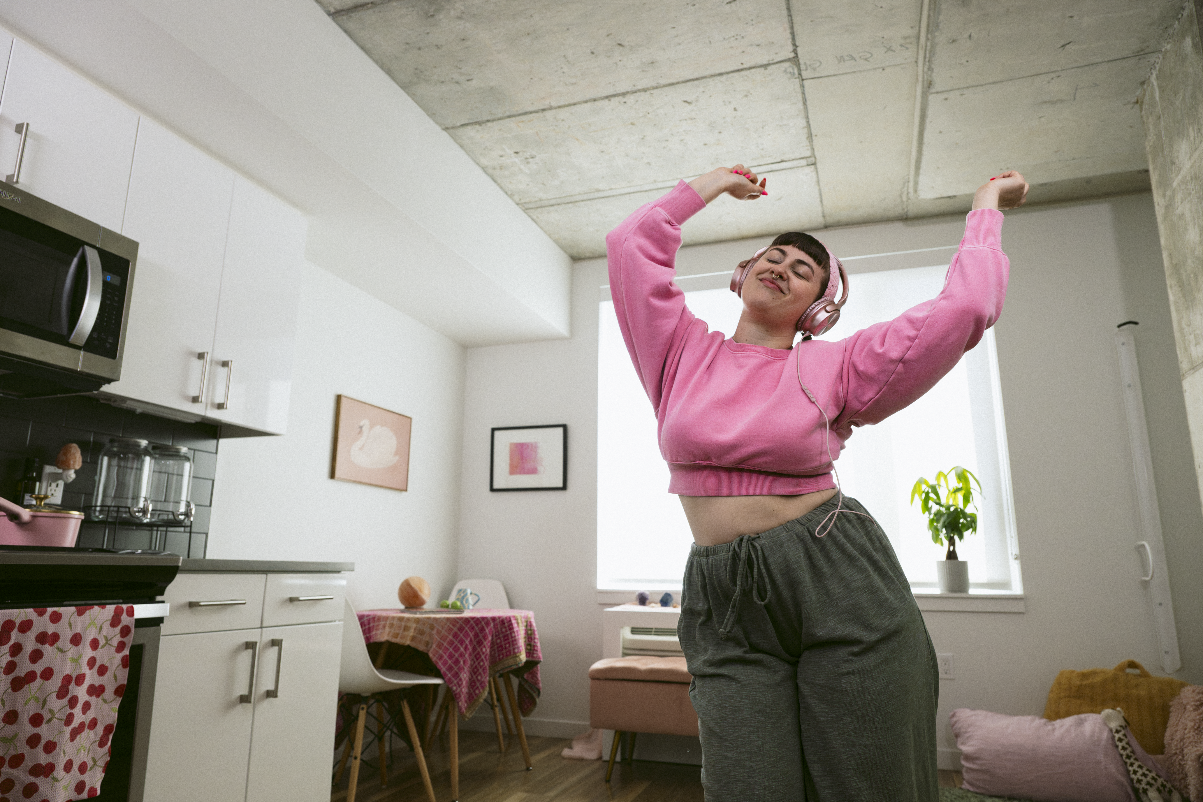 Woman wearing pink sweatshirt and pants dancing in her living room wearing headphones.